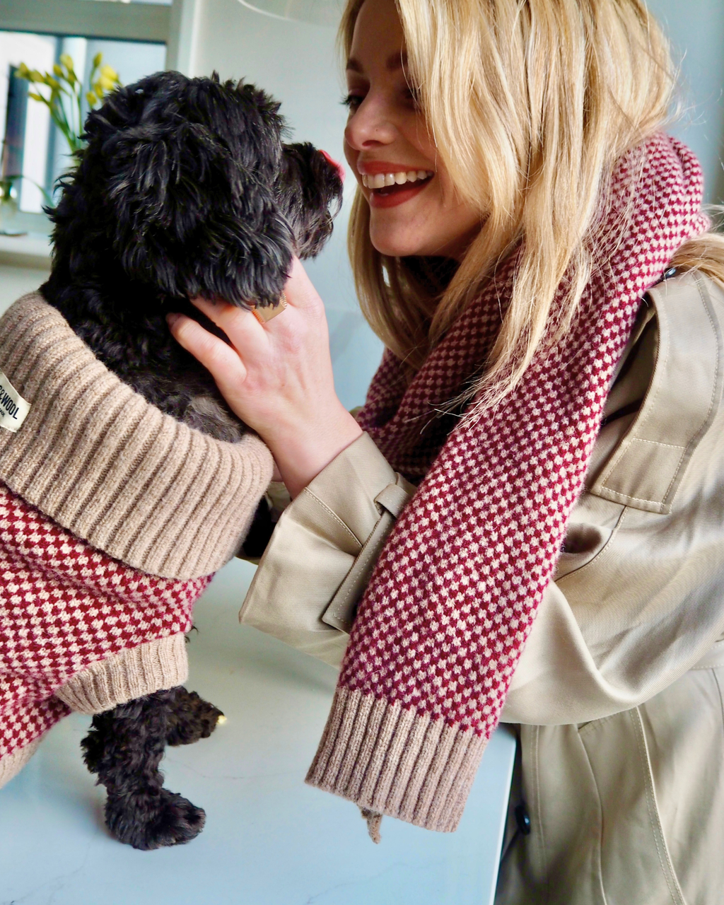 Woman in a burgundy check scarf cuddling a black dog in a matching jumper