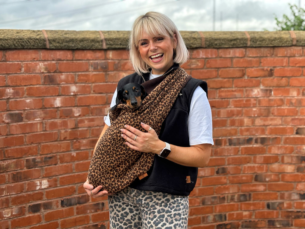 Smiling woman by a brick wall with a dachshund in a leopard Patterned Fleece Dog Sling