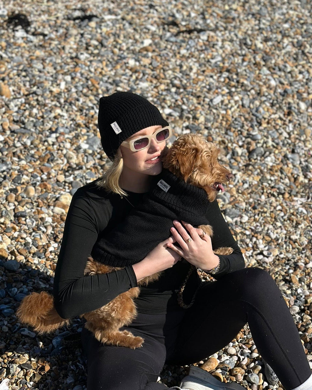 Woman in black hugging a golden dog in a black wool jumper on a pebble beach