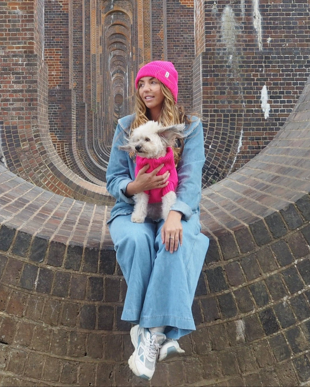 Woman in a pink beanie holding a small white dog in a pink wool jumper
