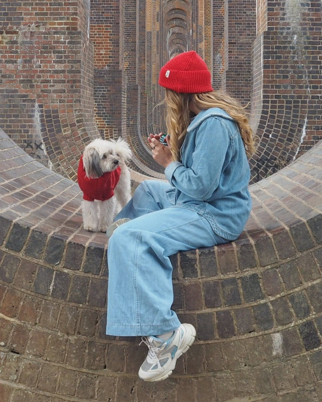 Woman in a red beanie with a small white dog in a red wool jumper on a brick wall