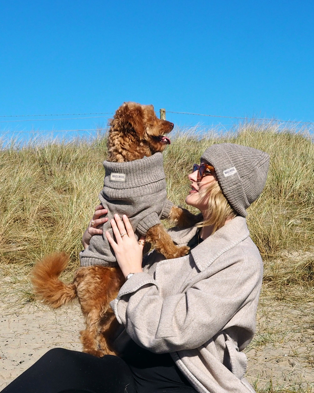 Woman in a beanie holding up a golden dog in a grey wool jumper in the dunes