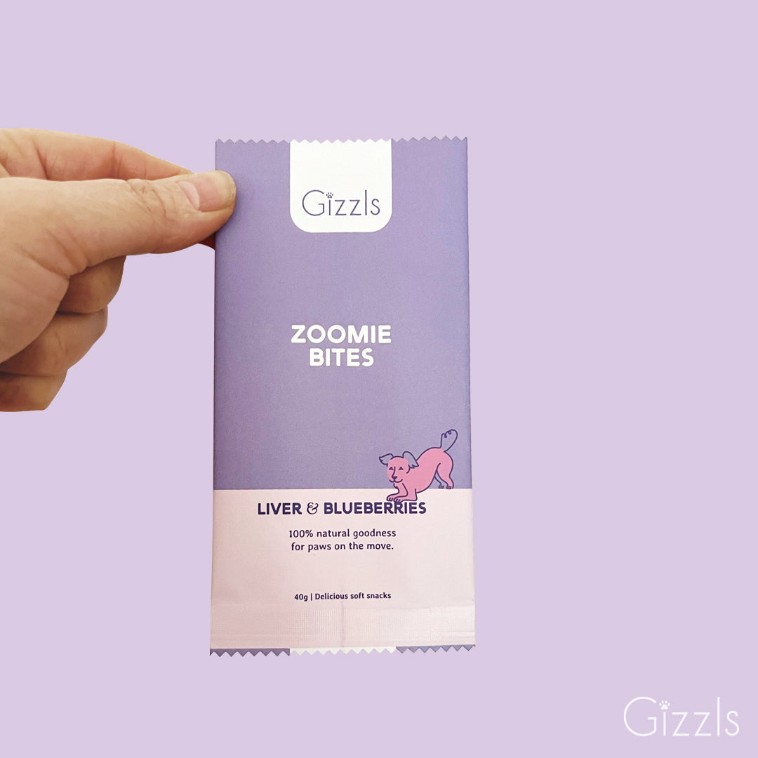 Hand holding a lilac Zoomie Bites Ostrich Liver & Blueberry snack pack on a lavender background