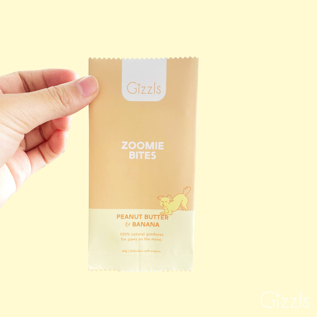 Hand holding a yellow Zoomie Bites Peanut Butter & Banana snack pack on a pale yellow background