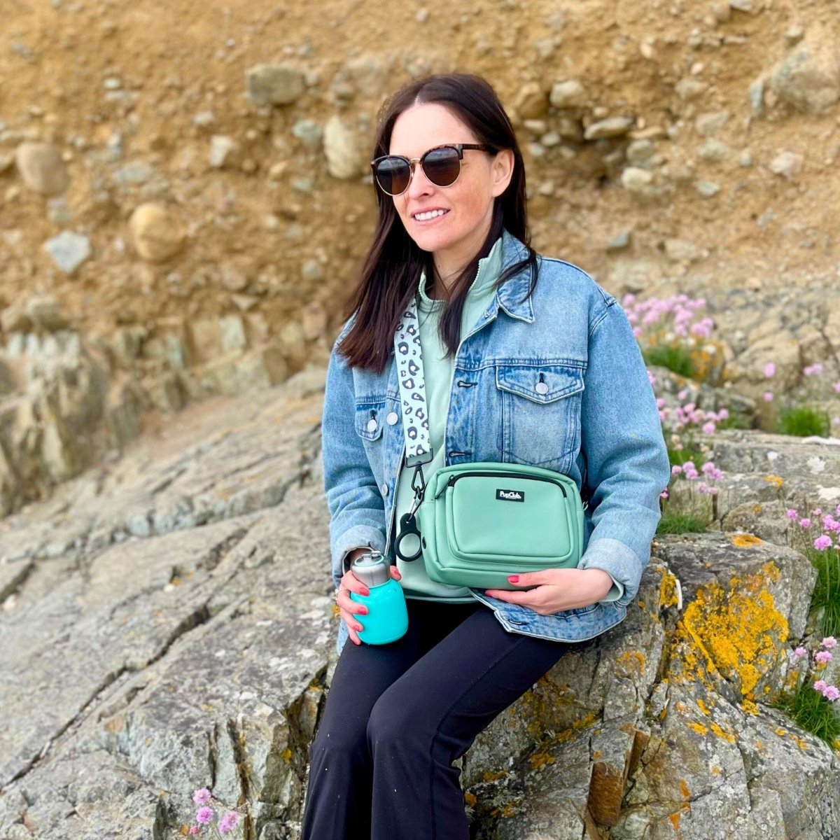 Woman sitting on rocks wearing a sage green crossbody dog walking bag