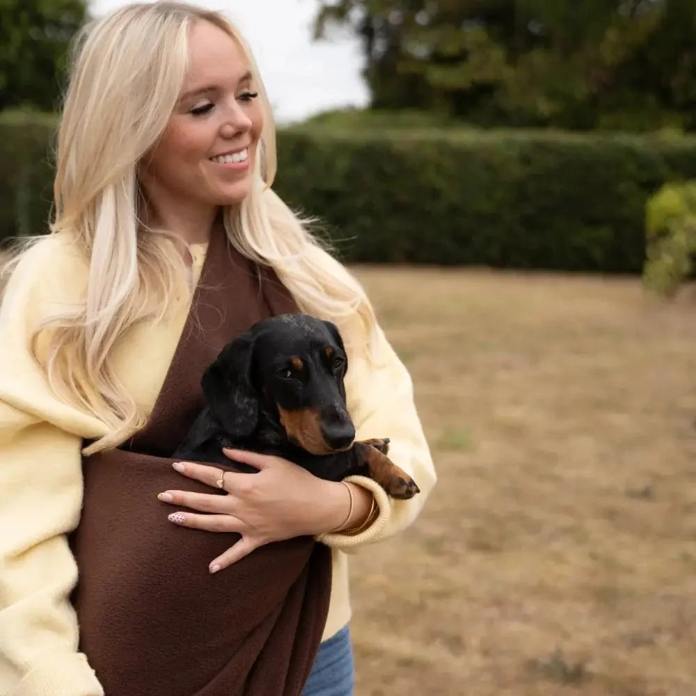 Smiling woman carrying a black dachshund in a chocolate Plain Fleece Dog Sling outdoors