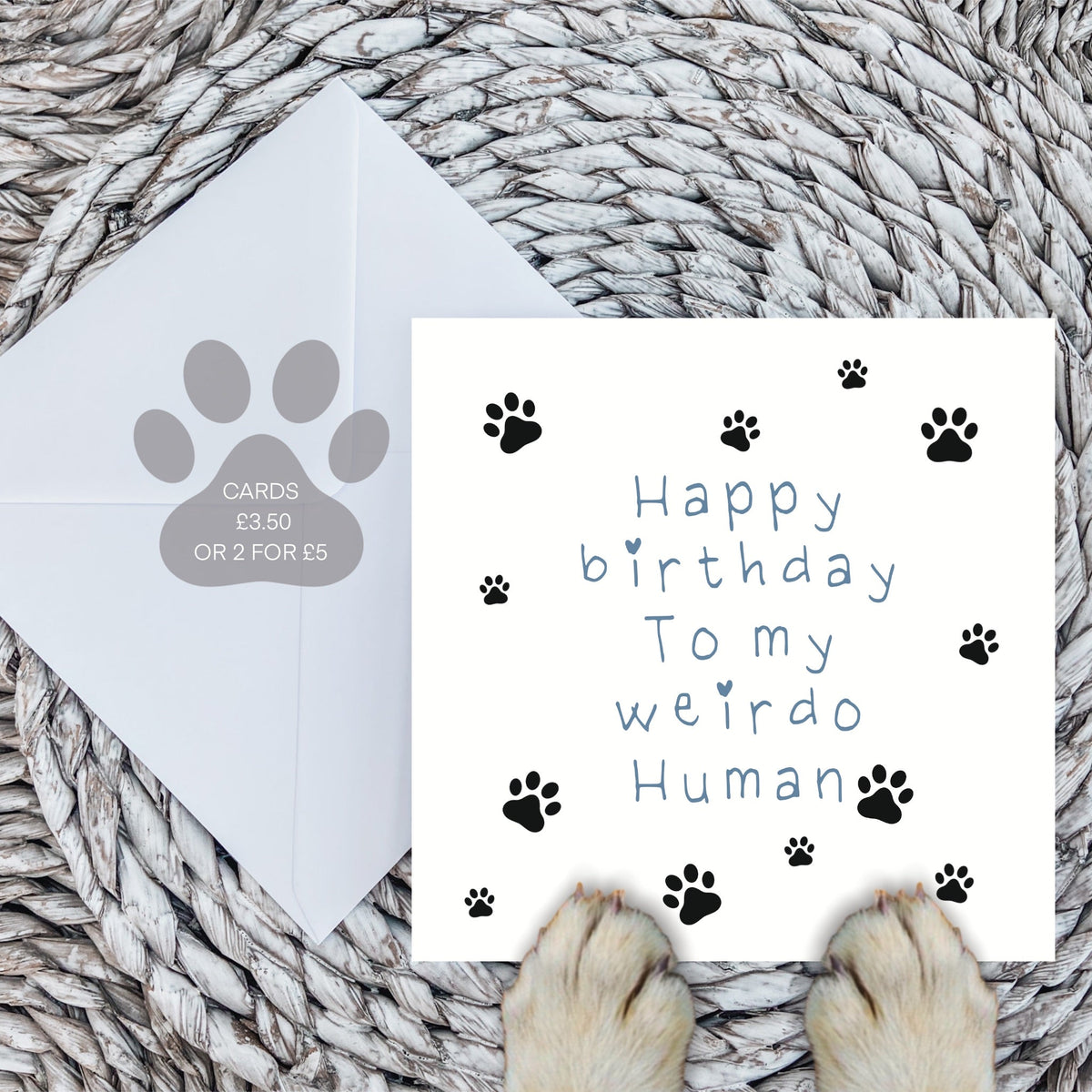 Blue weirdo human card and white envelope on a woven mat with dog paws