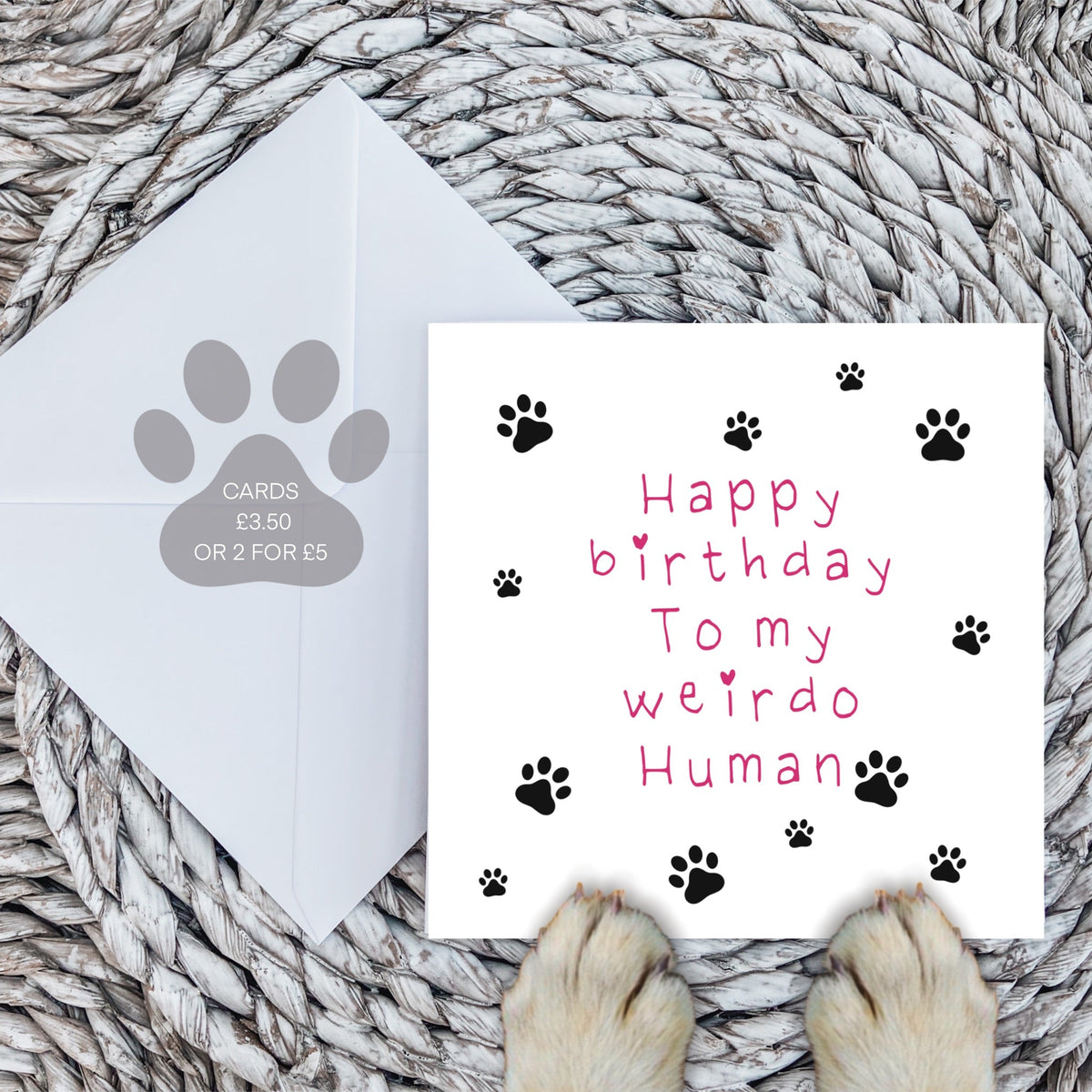 Pink weirdo human card and white envelope on a woven mat with dog paws