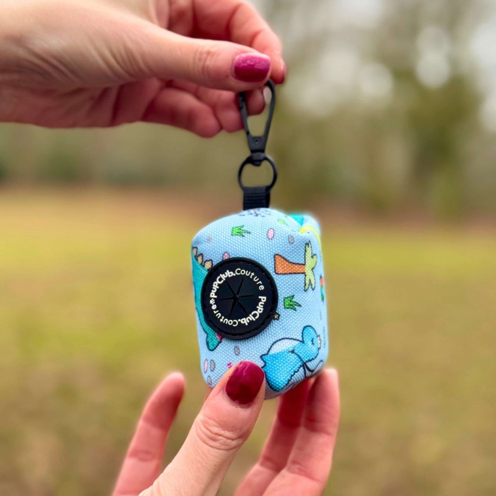 Hand holding the Jurassic Party Poo Bag Holder by its clip in a field