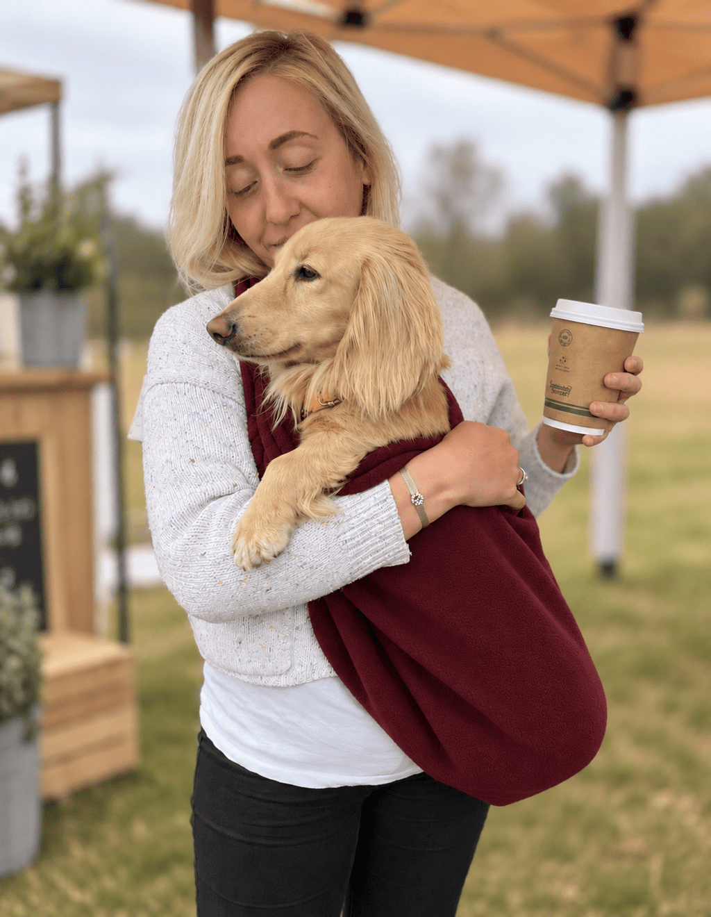 Woman at an outdoor event cuddling a cream dachshund in a wine Plain Fleece Dog Sling